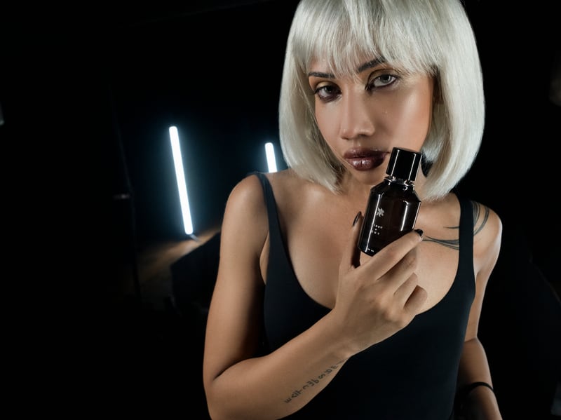 A woman in a white bob wig beside a standing neon tube, the flacon at her chest