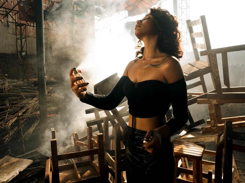 A woman seated among wooden chairs in smoke, the flacon in her hand