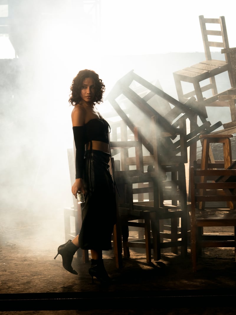 A woman standing full length in smoke beside a pile of wooden chairs, warm daylight behind her