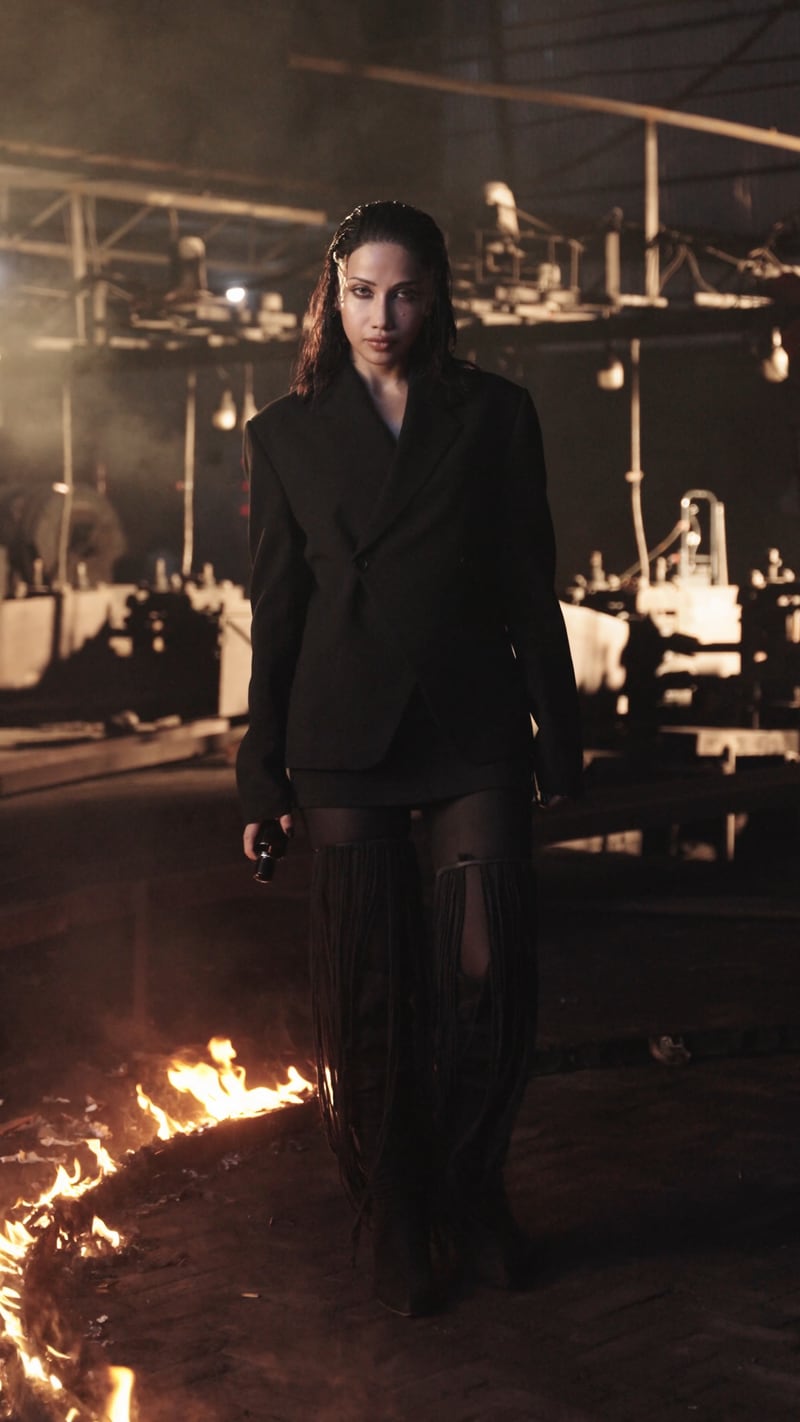 A woman walking towards camera in a machine hall, a ring of fire burning behind her